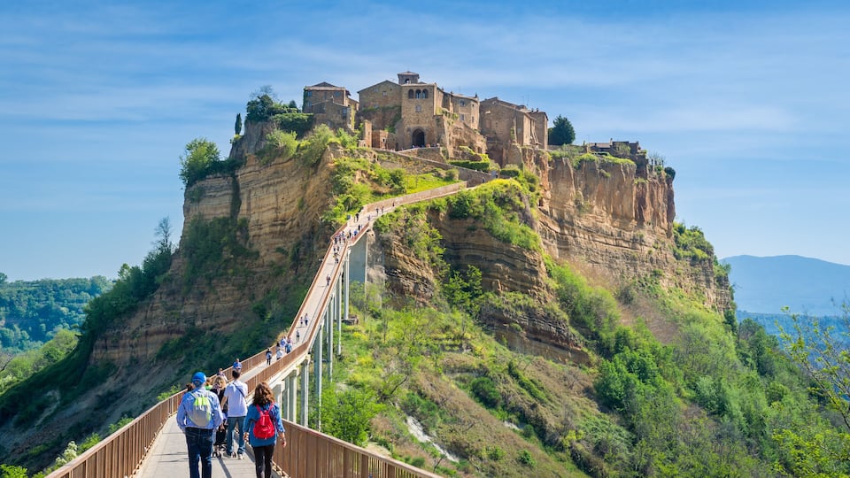A historic stone fortress on a cliff, a pedestrian bridge, and lush green hills.