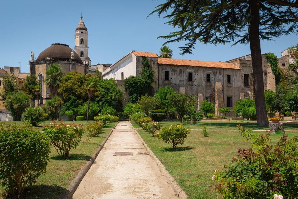 A historic building complex with a central dome and bell tower, surrounded by well-maintained gardens and pathways.