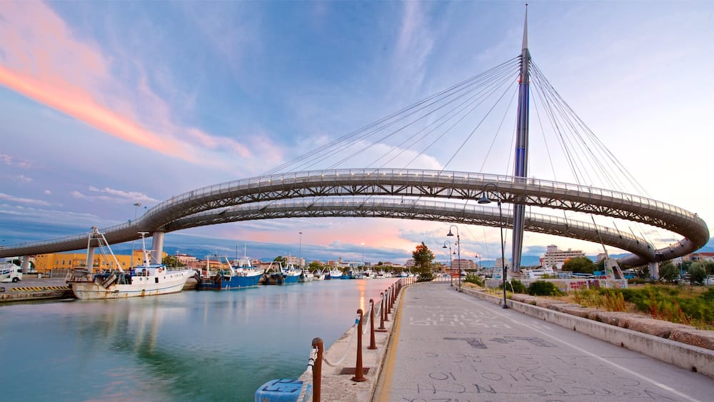A large, modern bridge spanning a body of water, with boats docked nearby and a cityscape in the background.