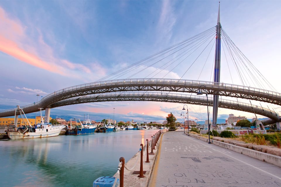 Uma grande ponte moderna atravessando um corpo d'água, com barcos atracados por perto e um horizonte urbano ao fundo.