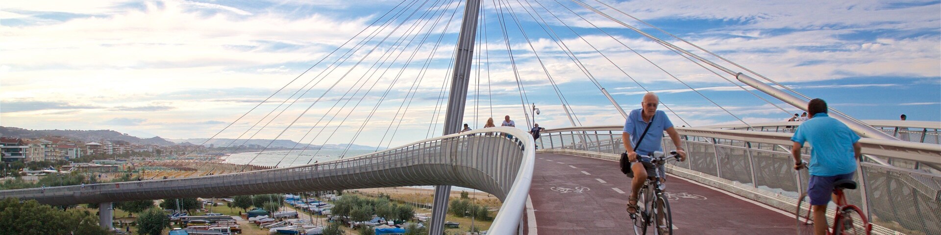 A modern cable-stayed bridge with a distinctive curved walkway, cyclists on the path, and a river below.