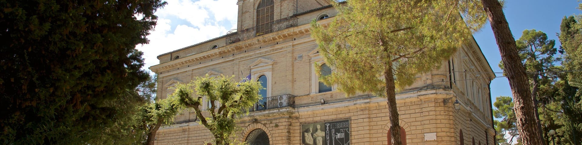 Un edificio histórico con ventanas de arco y un balcón, rodeado de árboles y un patio de grava.