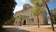 A historic building with arched windows and a balcony, surrounded by trees and a gravel courtyard.