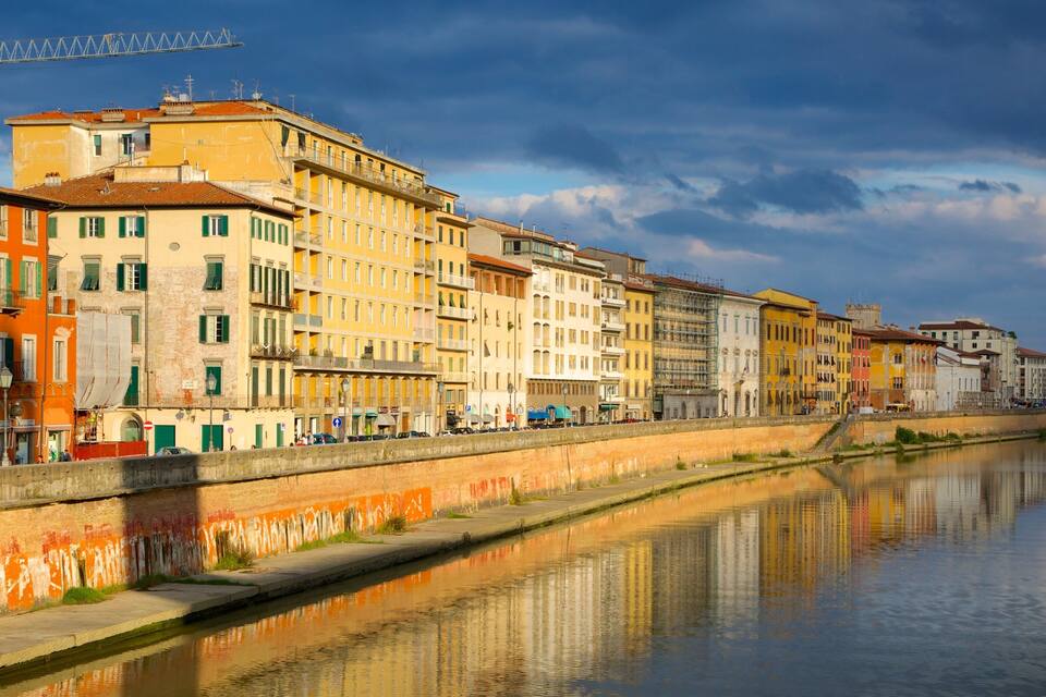 A canal with buildings reflecting on the water, a bridge in the background, and a partly cloudy sky.