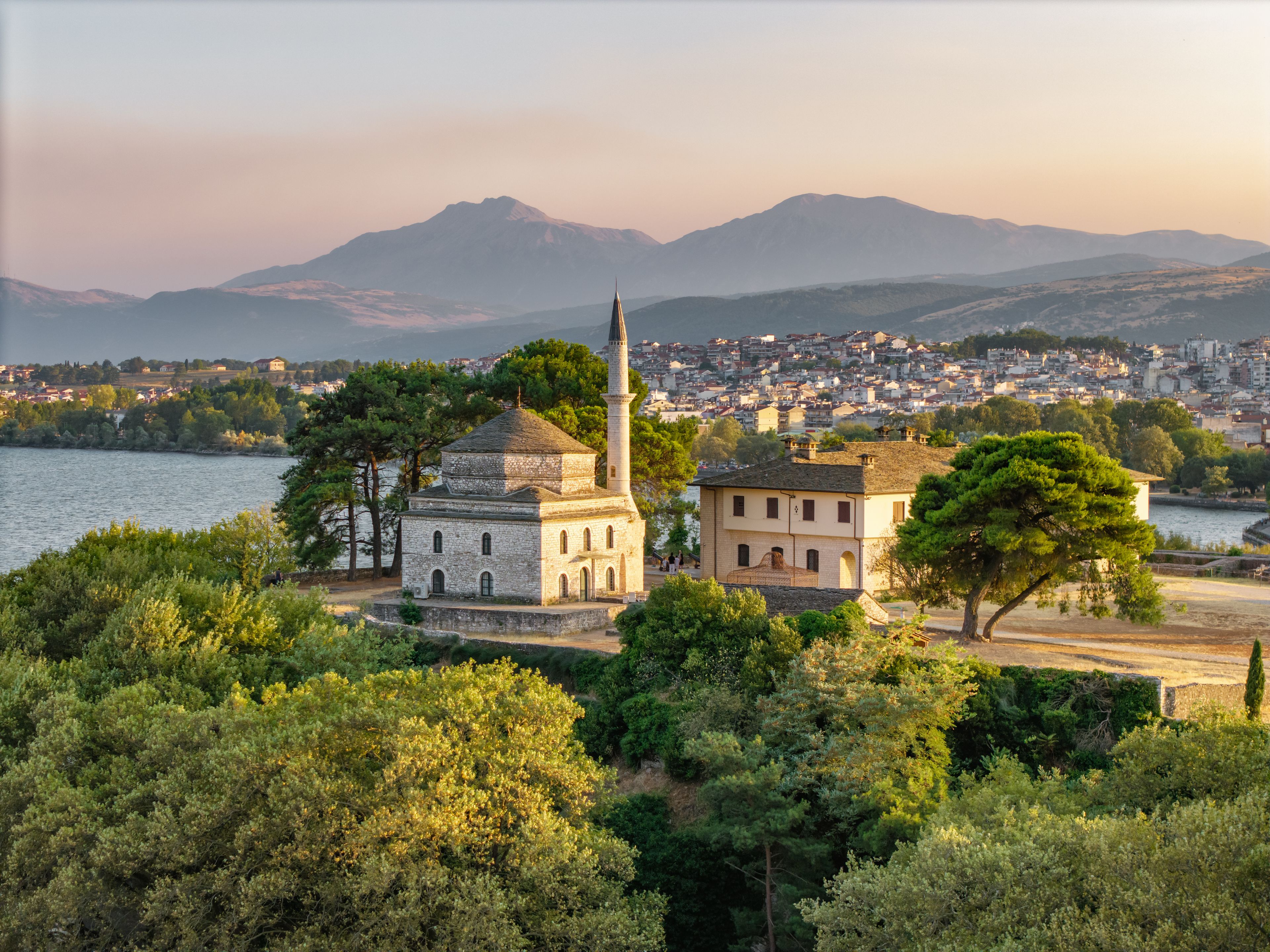 Un bâtiment historique doté d’une haute tour, entouré d’une végétation luxuriante et bordé d’un plan d’eau, avec une chaîne de montagnes en arrière-plan.