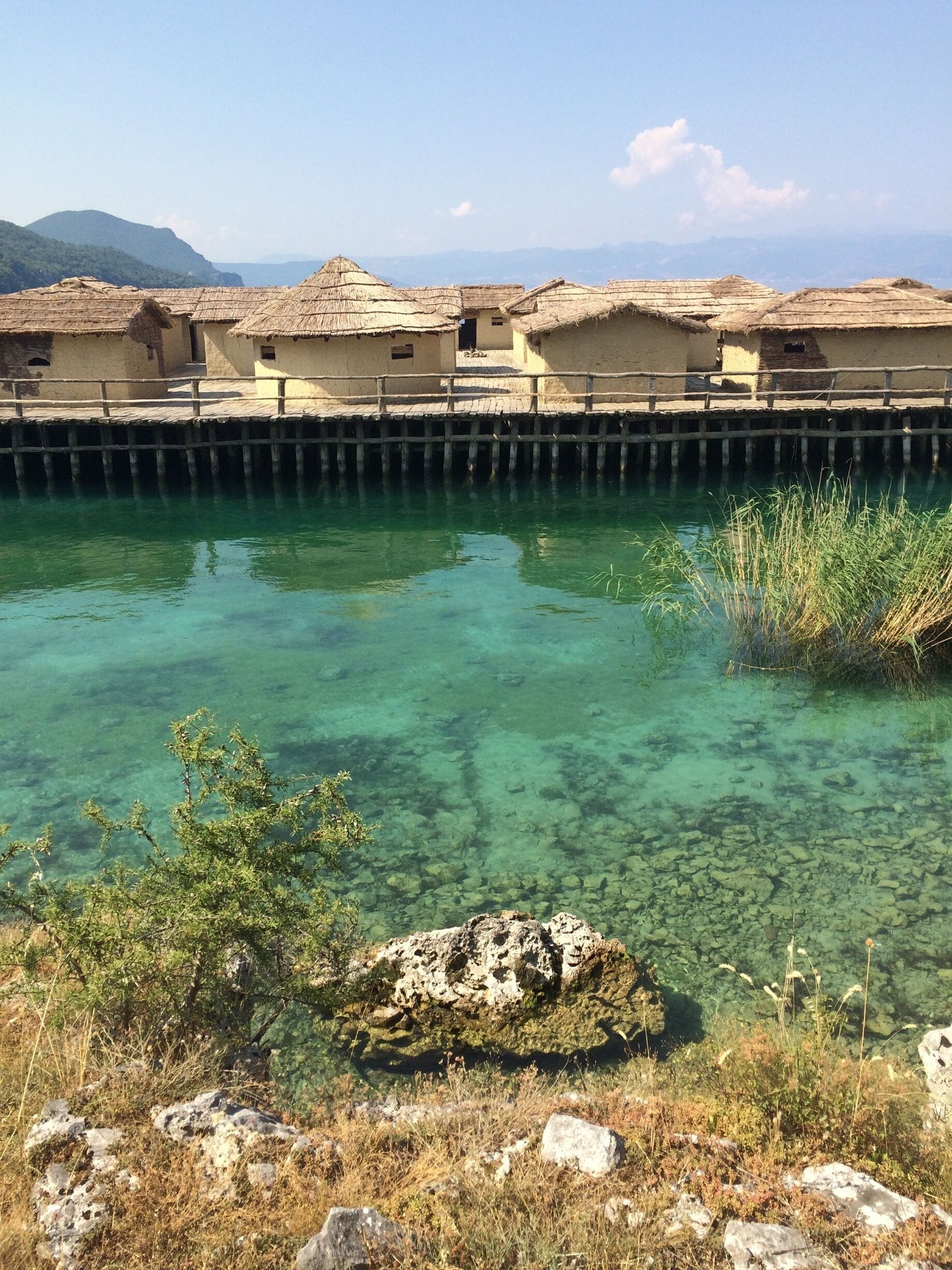 A clear water body with a wooden pier leading to traditional thatched-roof buildings.