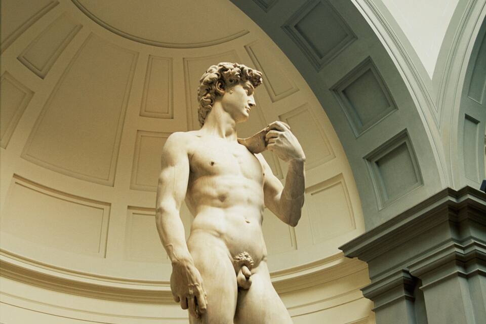 A marble statue of a standing man in a classical pose, set against a white wall with decorative molding and a coffered ceiling.