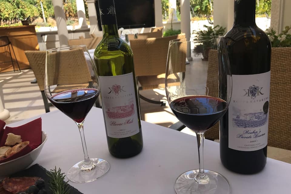 A table with two wine bottles and two wine glasses, surrounded by wicker chairs and a tablecloth.