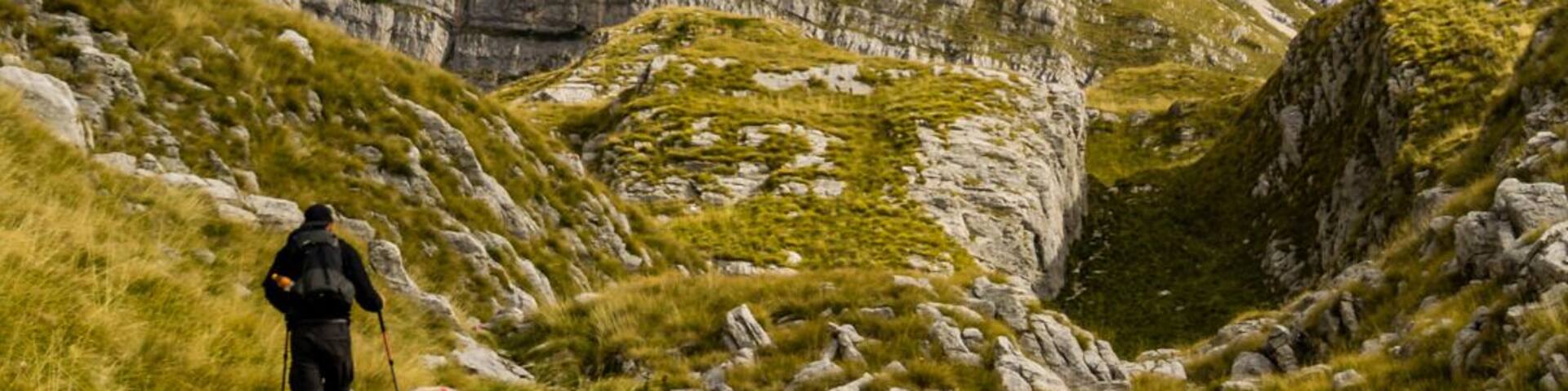 A hiker on a grassy mountain trail with rocky cliffs in the background.