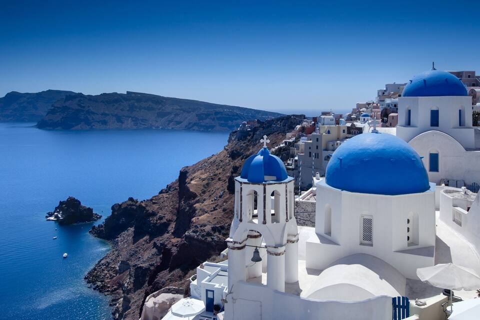 A coastal village with white buildings and blue domes, a rocky coastline, and a clear blue sea.