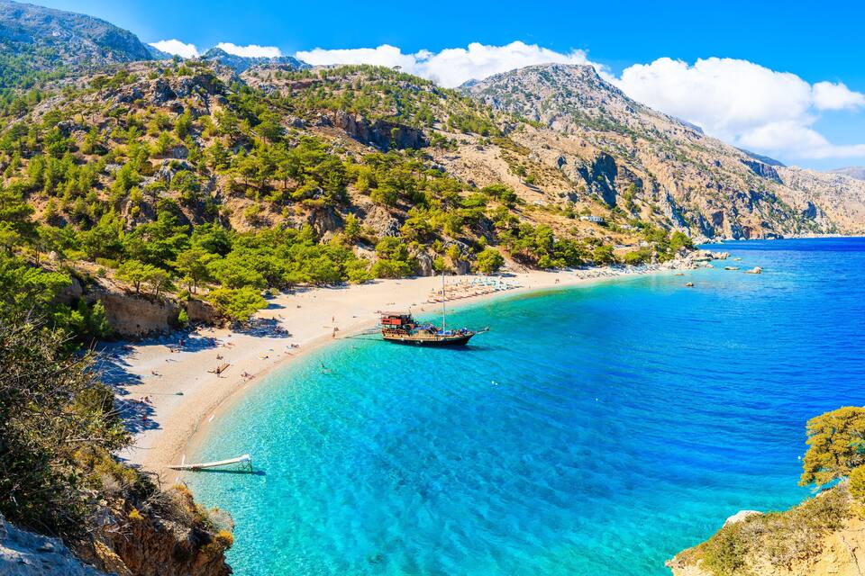A coastal landscape with a clear blue sea, a sandy beach, and a mountainous backdrop.