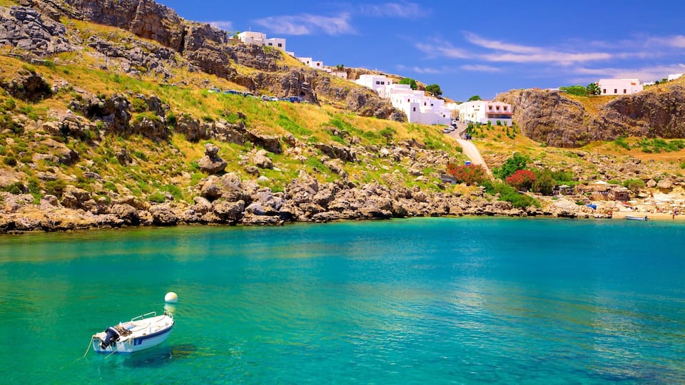 A coastal village with white buildings, rocky hills, and a boat in turquoise waters.