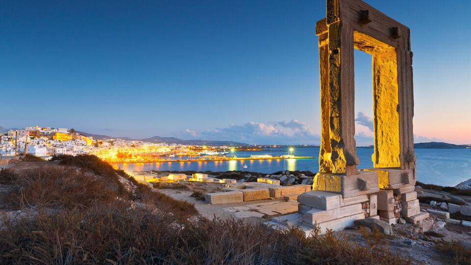 Une ville côtière au crépuscule, avec une imposante structure de pierre antique au premier plan et un port illuminé en arrière-plan.