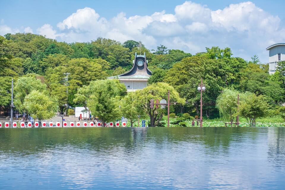 Um pagode japonês tradicional com uma torre de relógio, cercado por vegetação exuberante e um lago tranquilo.