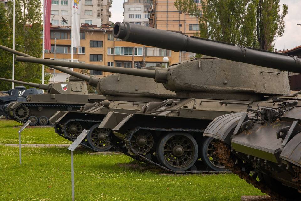 A military tank on display with a large gun, surrounded by grass and buildings in the background.