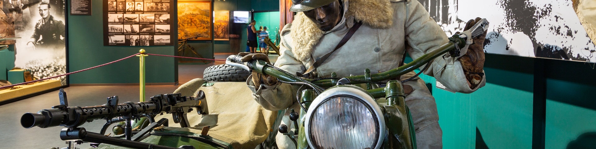 Uma motocicleta militar antiga com uma metralhadora montada, um capacete com óculos de proteção, e uma exposição de fotografias e documentos históricos ao fundo.