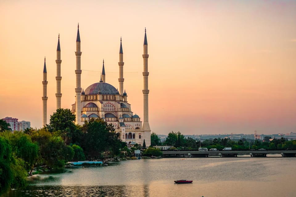 A mosque with multiple minarets and a dome, situated by a river with a bridge in the background.