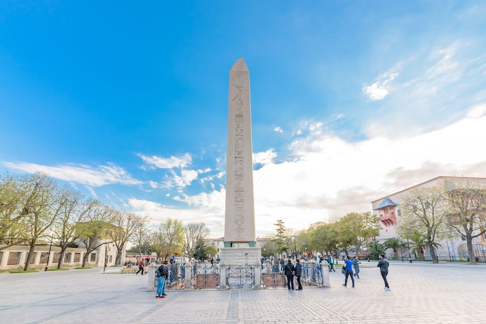 A tall obelisk monument in a public square with people walking around.