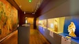 A museum corridor with wooden floors, illuminated exhibits, and a large mural on the left wall.