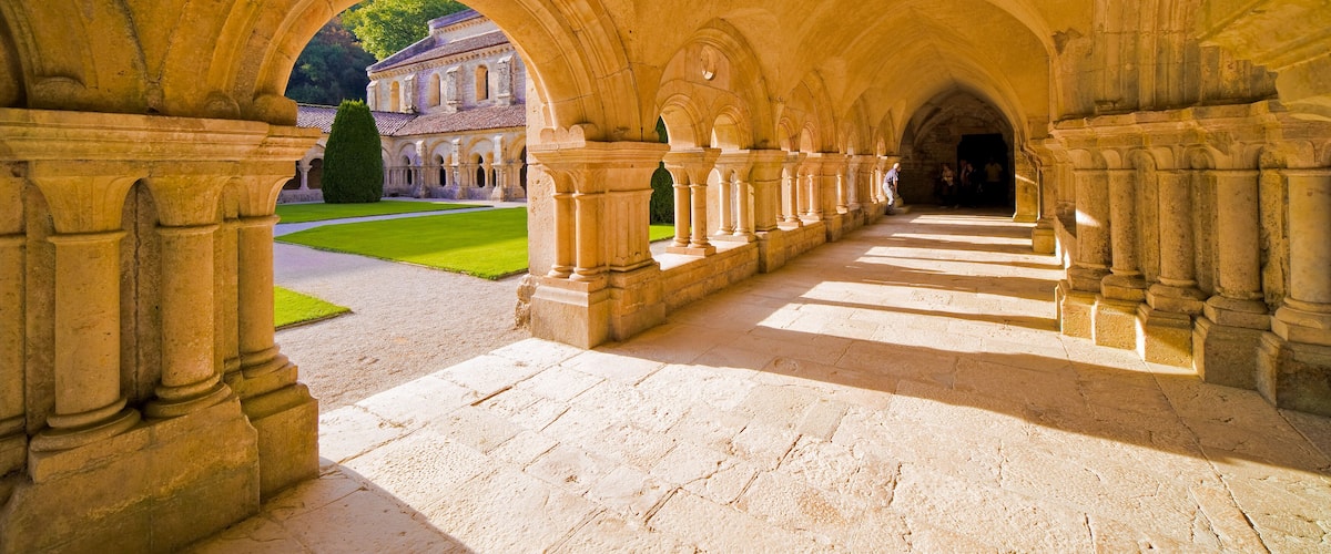 An arched corridor with stone columns, leading to a building with a green lawn and trees.