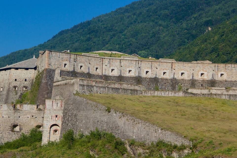 A historic stone fortress with multiple towers and a large wall, surrounded by a grassy hill and dense forest.