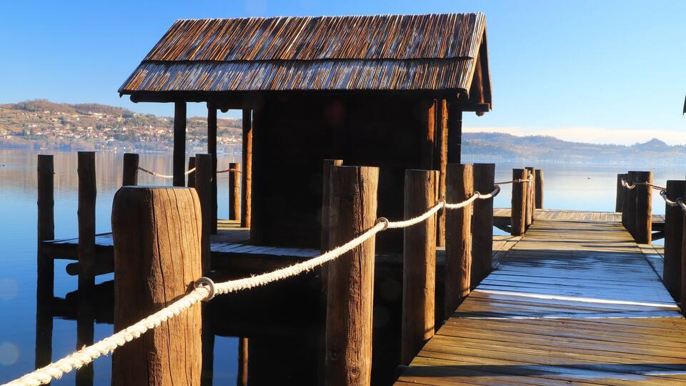 A wooden dock with a small hut at the end, leading into calm water with a distant shoreline.