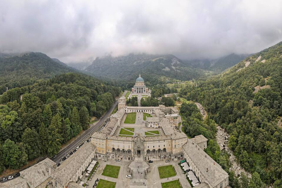 Vista aérea de un complejo de edificios históricos rodeado de verdes colinas y montañas, bajo un cielo despejado.