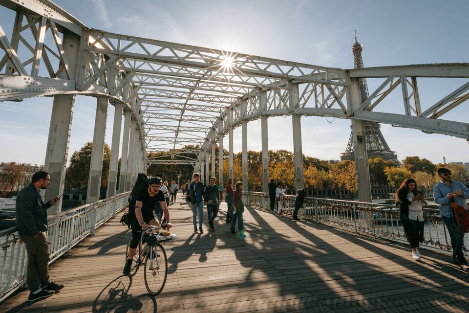 A bridge with a metal framework, people walking, and the Eiffel Tower in the background.