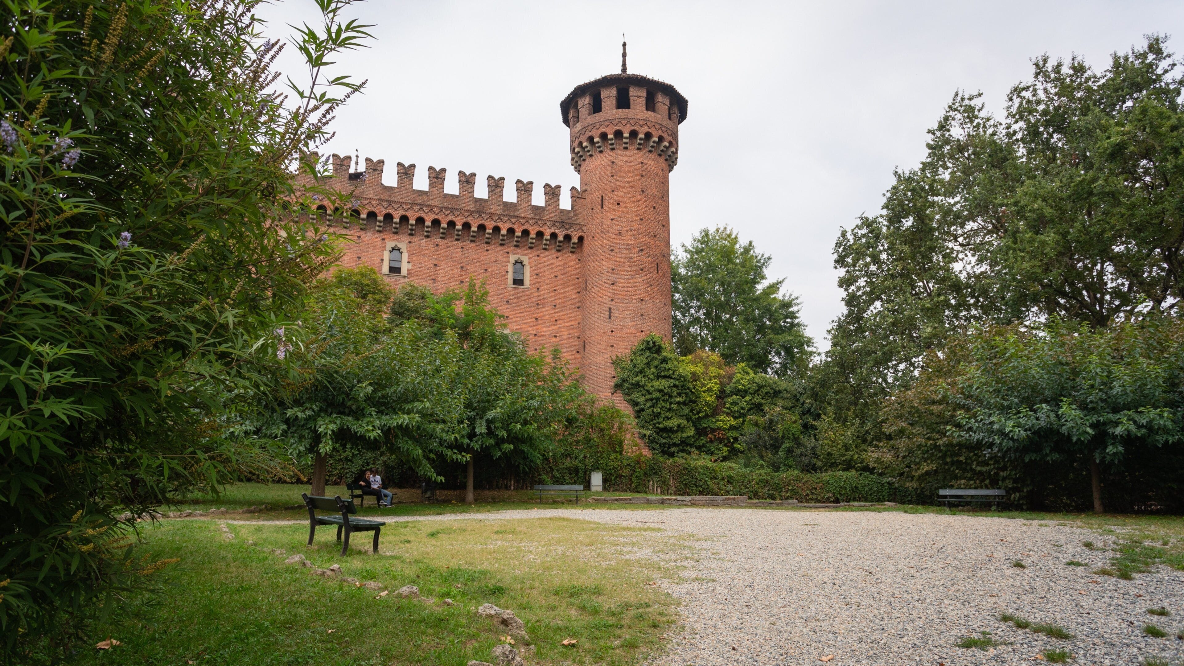 Ett slott med ett högt torn, omgivet av frodig grönska och en grusgång med bänkar.