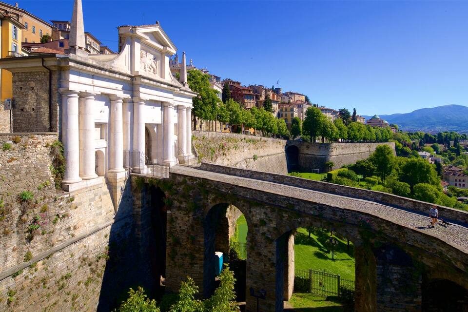 Un puente de piedra con arcos, un edificio clásico blanco con columnas y un paisaje urbano con edificios y vegetación al fondo.