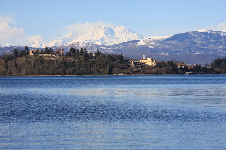 Um lago com uma igreja no topo de uma colina, montanhas nevadas ao fundo e um céu claro.
