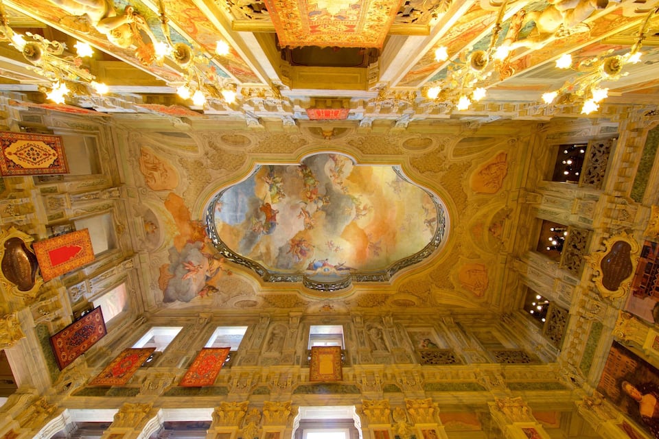 Una stanza sfarzosa con affreschi elaborati sul soffitto, lampadari dorati e decorazioni murali intricate.