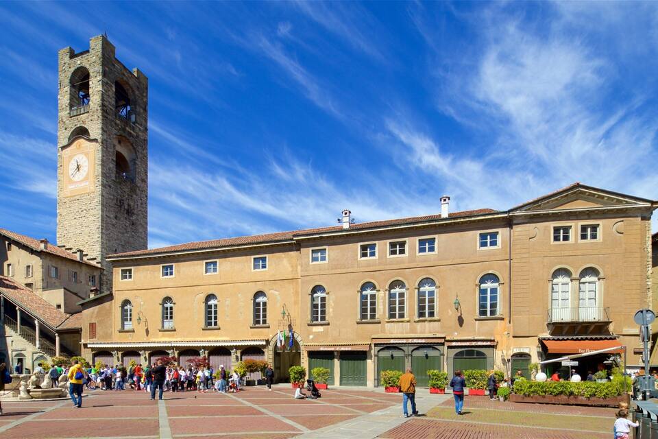 A historic square with a prominent clock tower, a large building with arched windows, and people walking around.