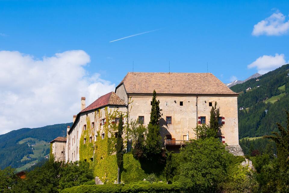 Ein historisches Gebäude mit rotem Ziegeldach, umgeben von üppiger Vegetation und Bergen im Hintergrund.