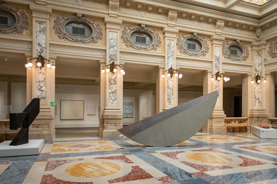 A grand hall with ornate columns, a large abstract sculpture, and a patterned floor.