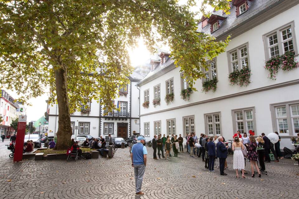 A cobblestone square with a large tree, white buildings with flower boxes, and people gathered.