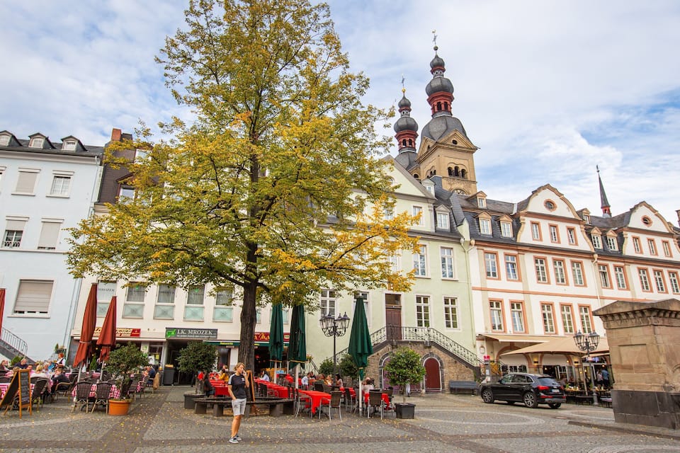 Una plaza adoquinada con mesas al aire libre, un gran árbol y edificios históricos con una torre de iglesia.
