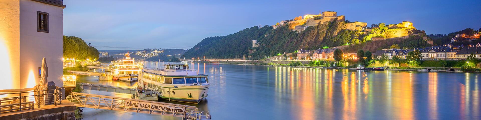 A calm river with boats docked, a castle on a hill, and a building with a blue roof.