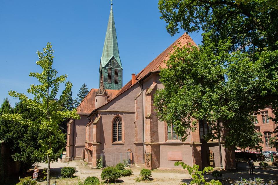 Een historische kerk met een hoge spits en een rode pannendak, omgeven door weelderige groene bomen.