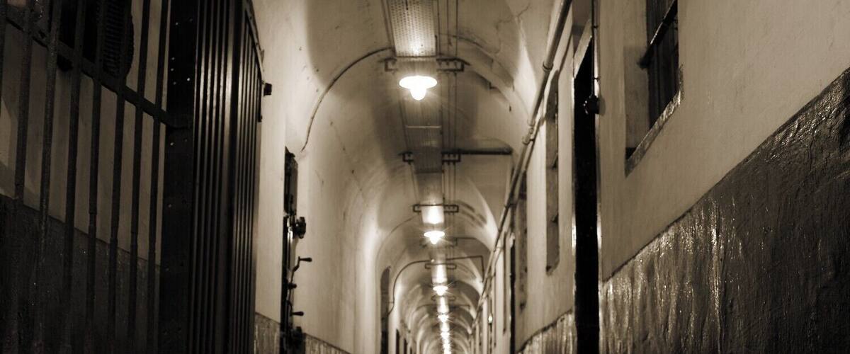 A long, dimly lit corridor with metal bars on one side and a concrete floor, leading to a light at the end.