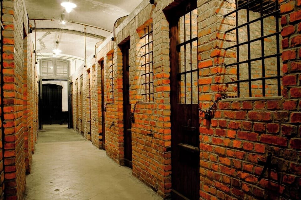A brick corridor with barred windows and doors, leading to a white door at the end.