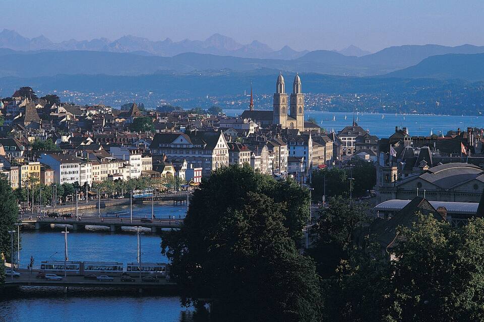 Stadsgezicht met een rivier, historische gebouwen en bergen op de achtergrond.