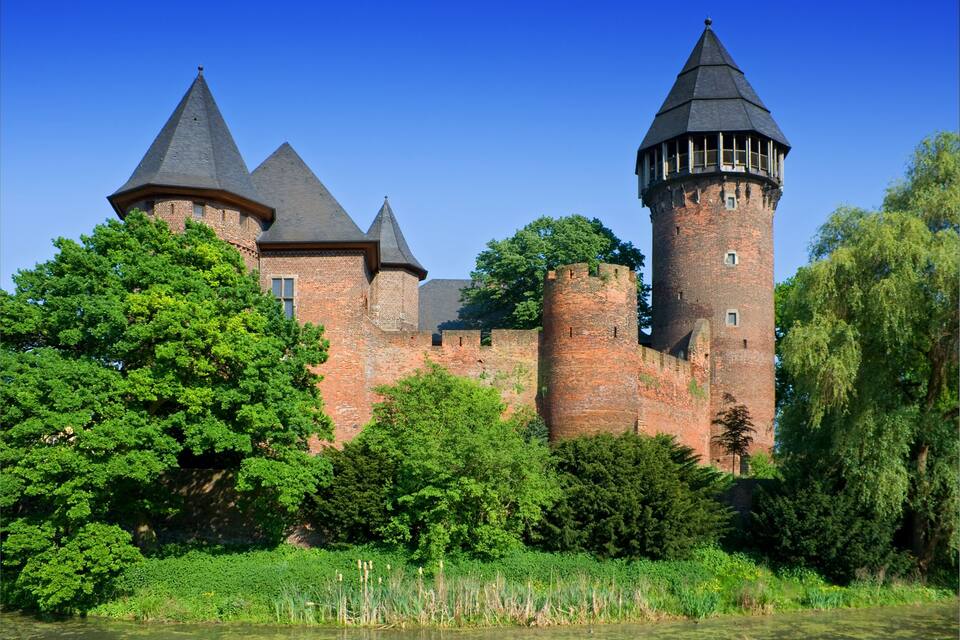 A historic castle with multiple towers, surrounded by lush greenery and a river.