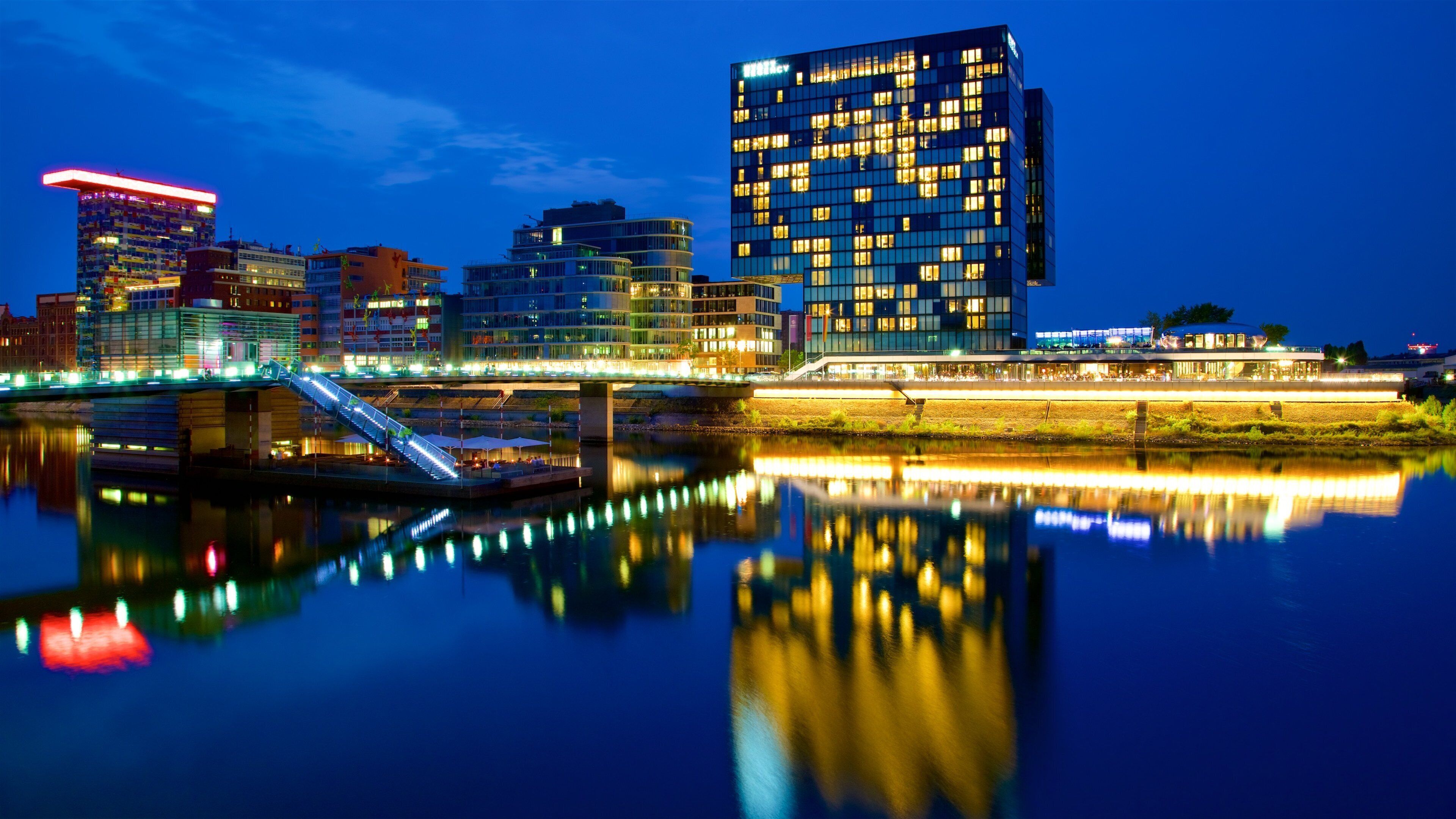 A nighttime cityscape with illuminated buildings reflecting on a river.