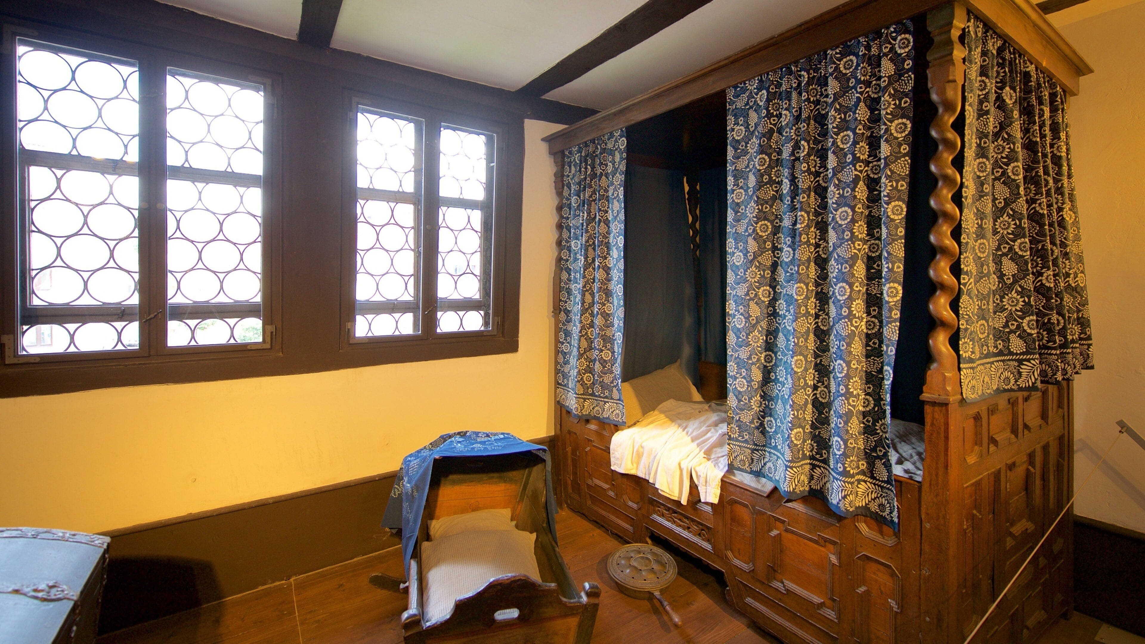 Una cama de madera tradicional con dosel, un taburete pequeño y una ventana con diseño geométrico.