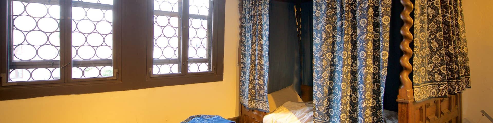 Una cama de madera tradicional con dosel, un taburete pequeño y una ventana con diseño geométrico.