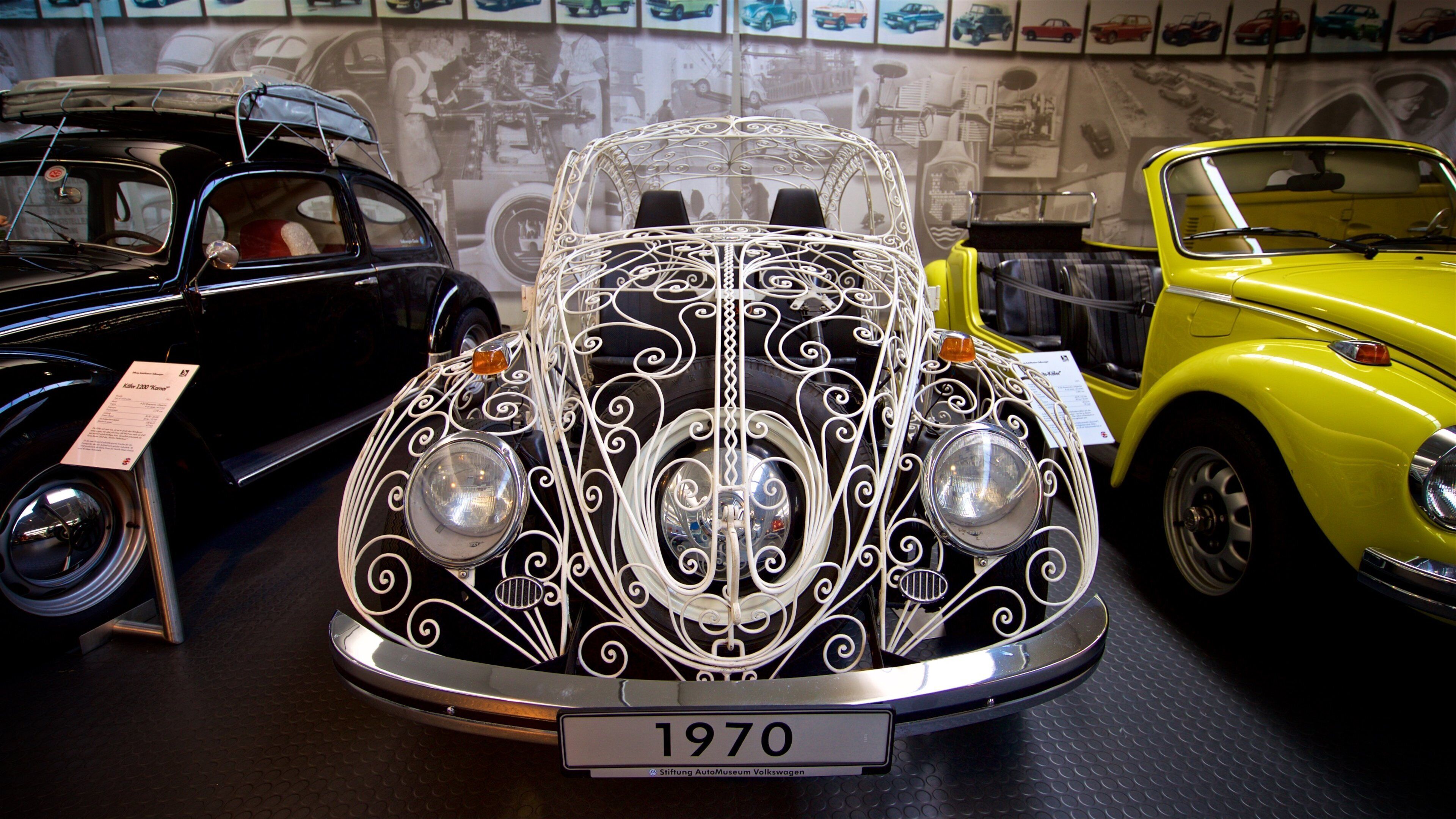 A vintage car with intricate white designs, labeled '1970', in a museum setting with other classic cars and a wall of car images in the background.