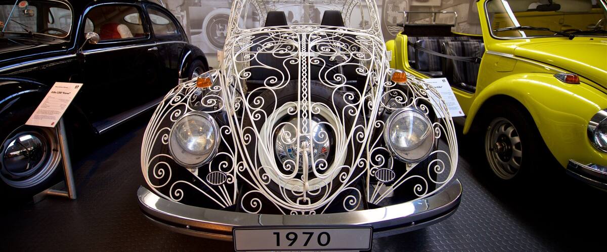 A vintage car with intricate white designs, labeled '1970', in a museum setting with other classic cars and a wall of car images in the background.