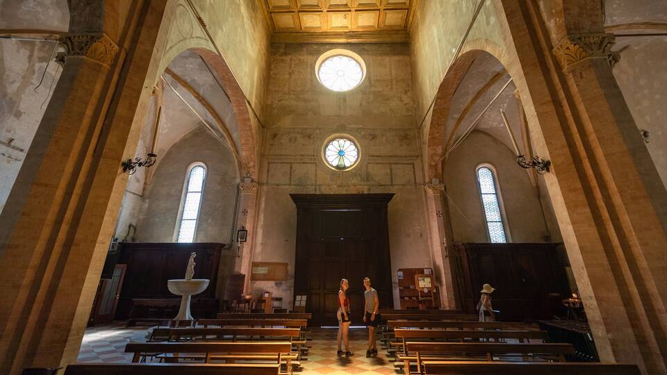 An interior of a church with arched ceilings, a central stained glass window, and wooden pews.