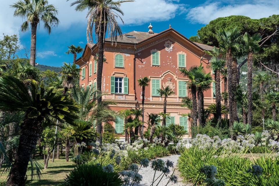 A historic building with green shutters, surrounded by palm trees and flowering plants.